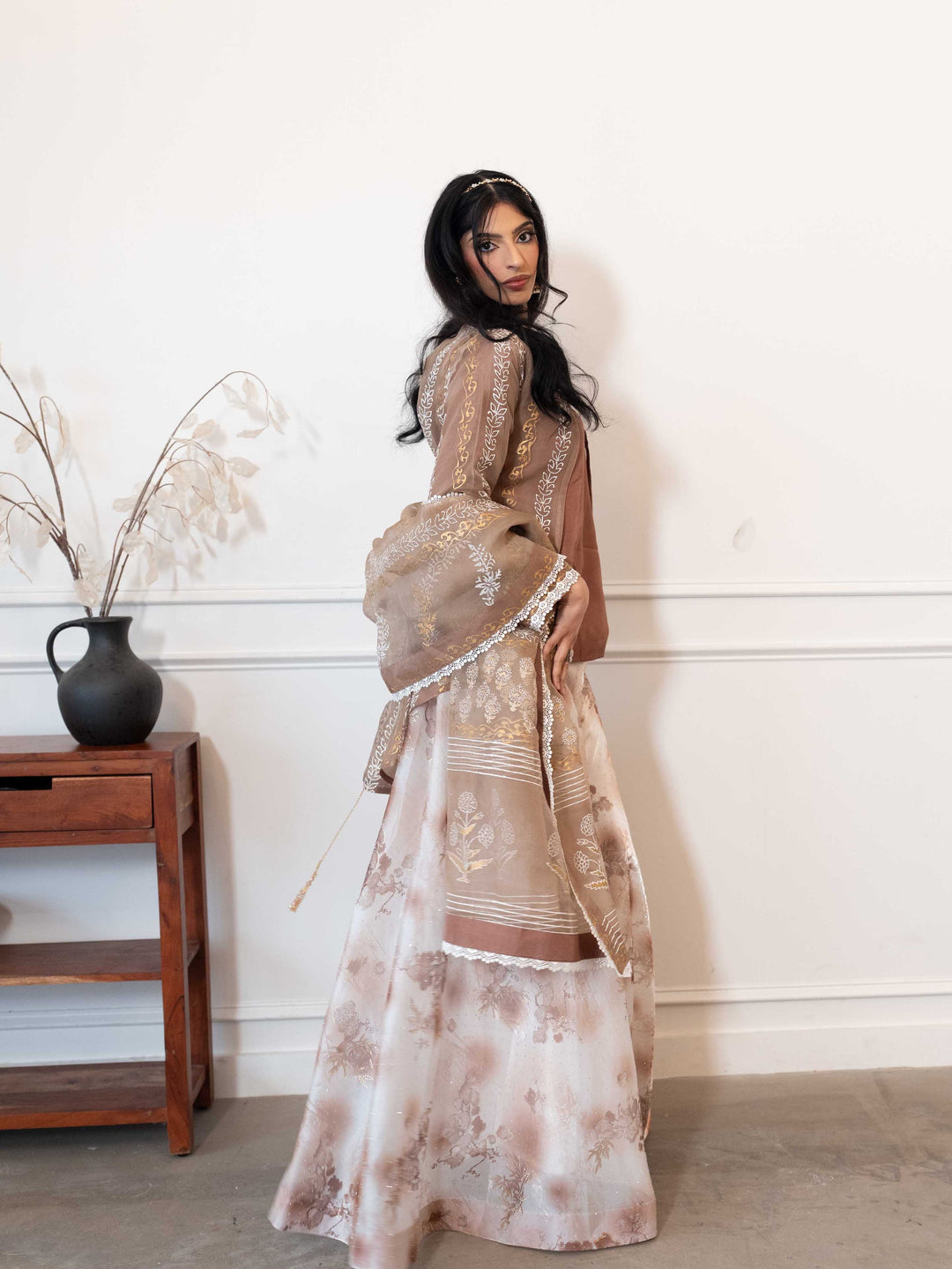 Woman in a floral outfit standing against a white wall with a wooden side table.